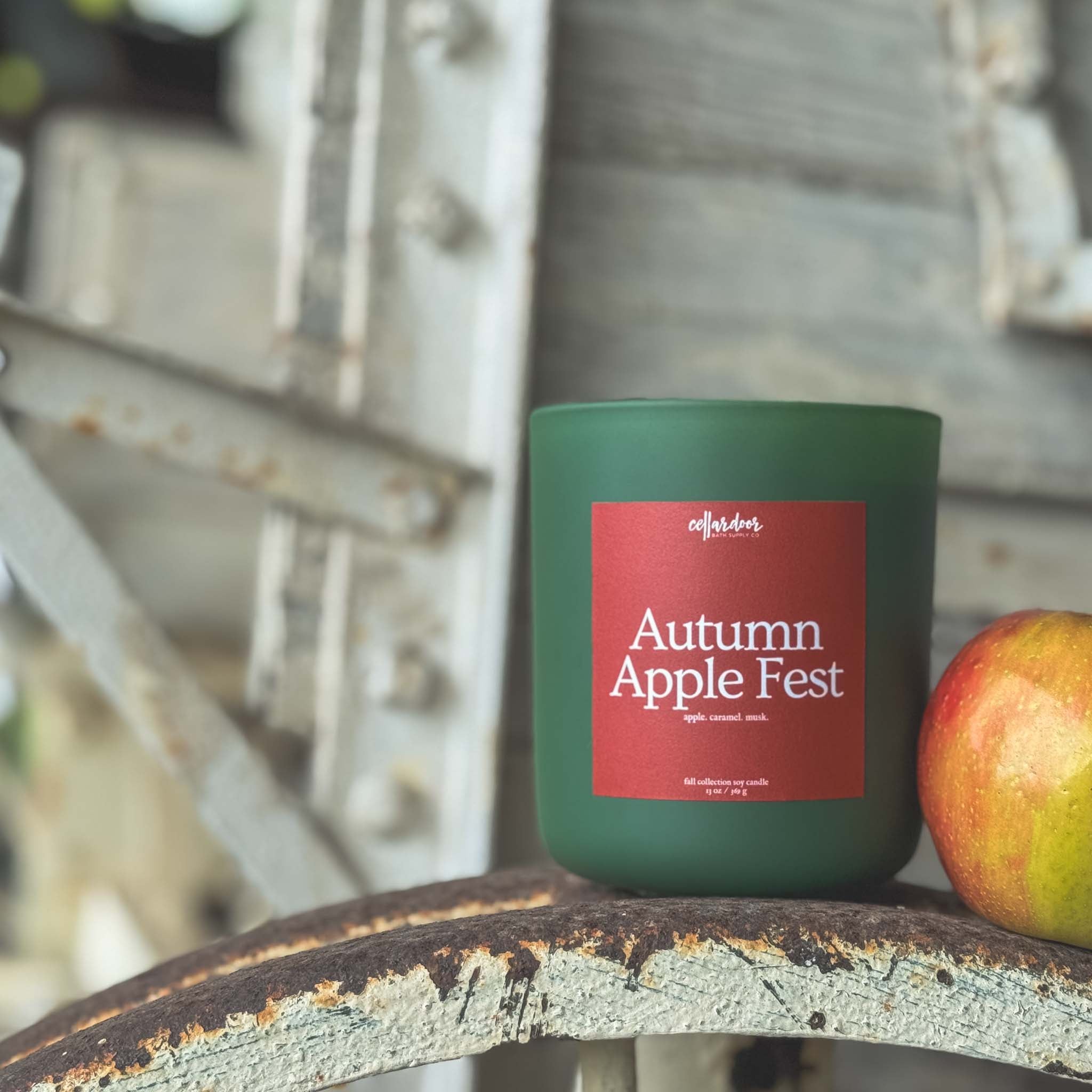 Candle labeled 'Autumn Apple Fest' on a metal surface with an apple beside it.