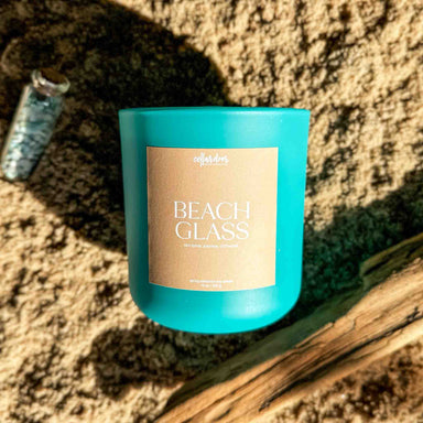 Blue candle labeled 'Beach Glass' on a sandy surface with driftwood.