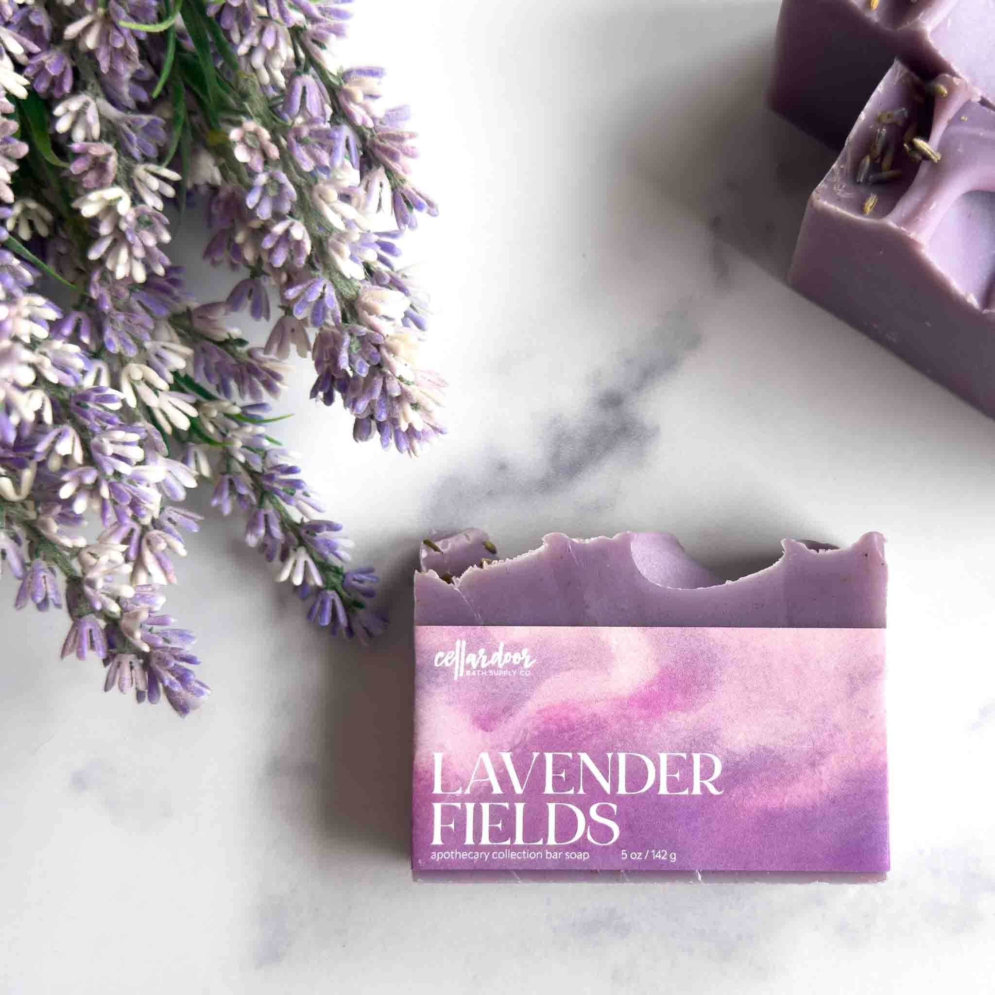Lavender Fields artisan soap by Cellar Gardens, shown in its purple paper packaging on a white marble surface with lavender flowers and a second unwrapped bar of soap nearby.