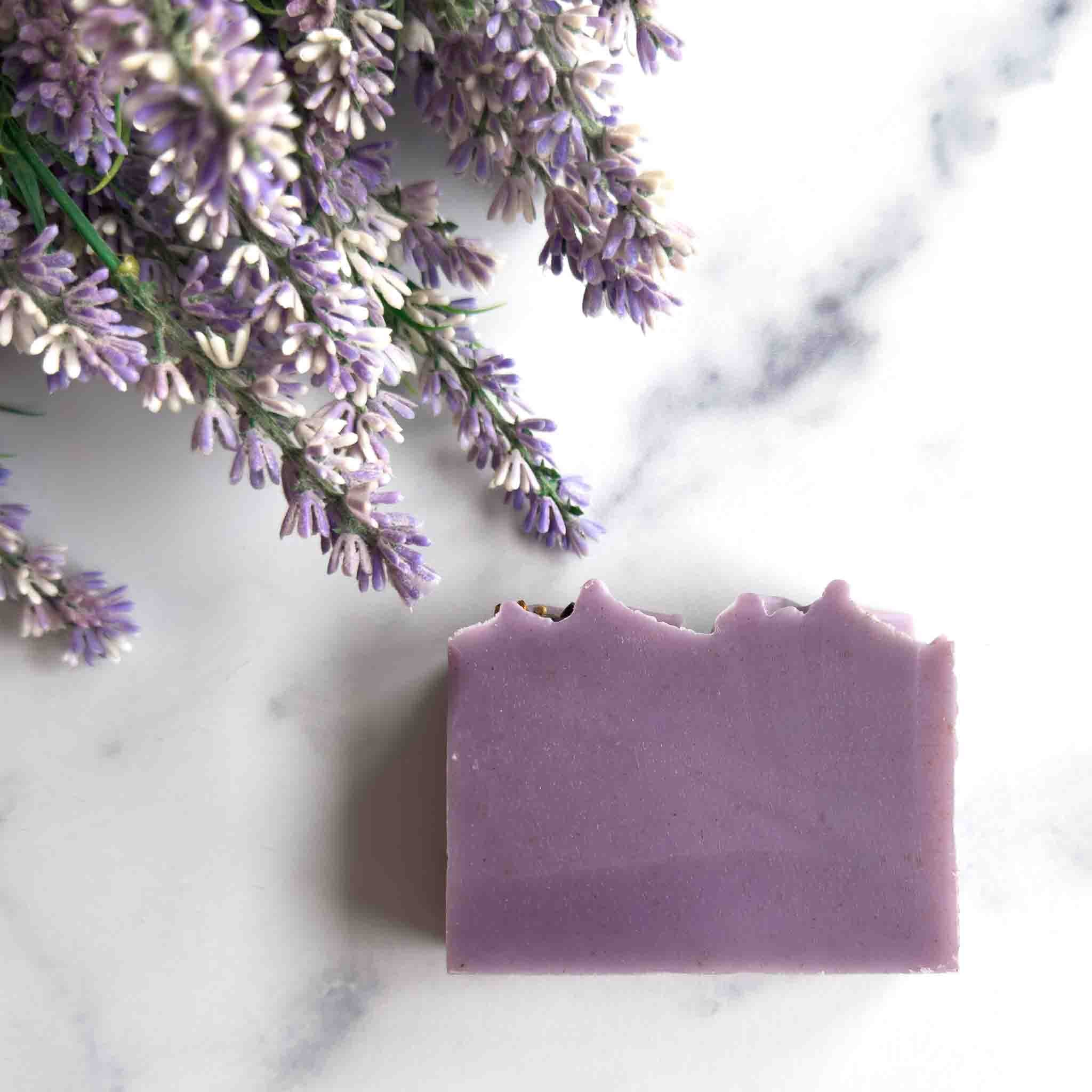 A rectangular bar of lavender-colored soap with rustic, unevenly broken edges, styled on a white marble surface next to dried lavender flowers.