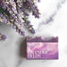 A bar of lavender soap labeled 'collaborative' and 'LAVENDER FIELDS' sits on a white marble surface next to dried lavender sprigs.
