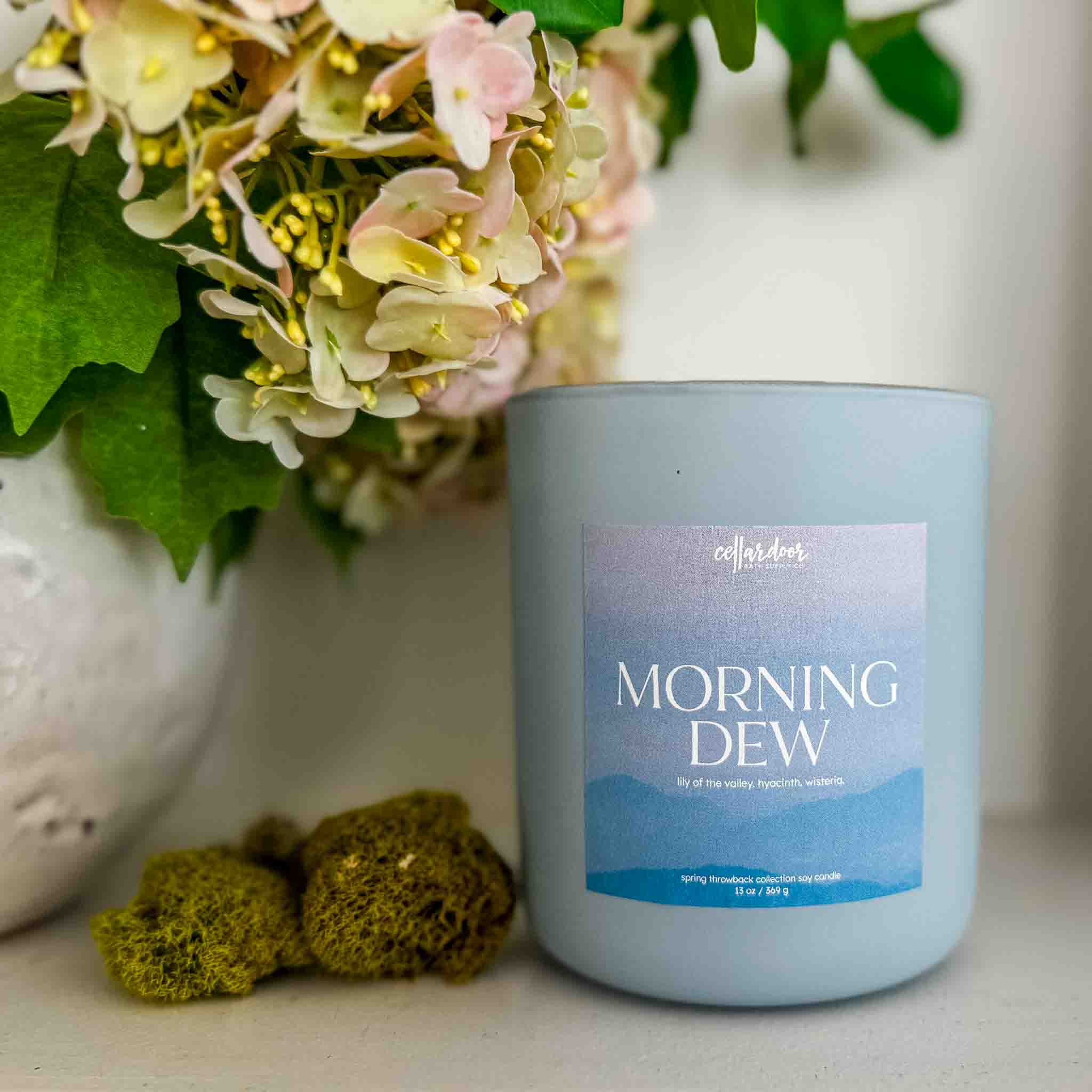 Candle labeled 'Morning Dew' with flowers and decorative elements in the background