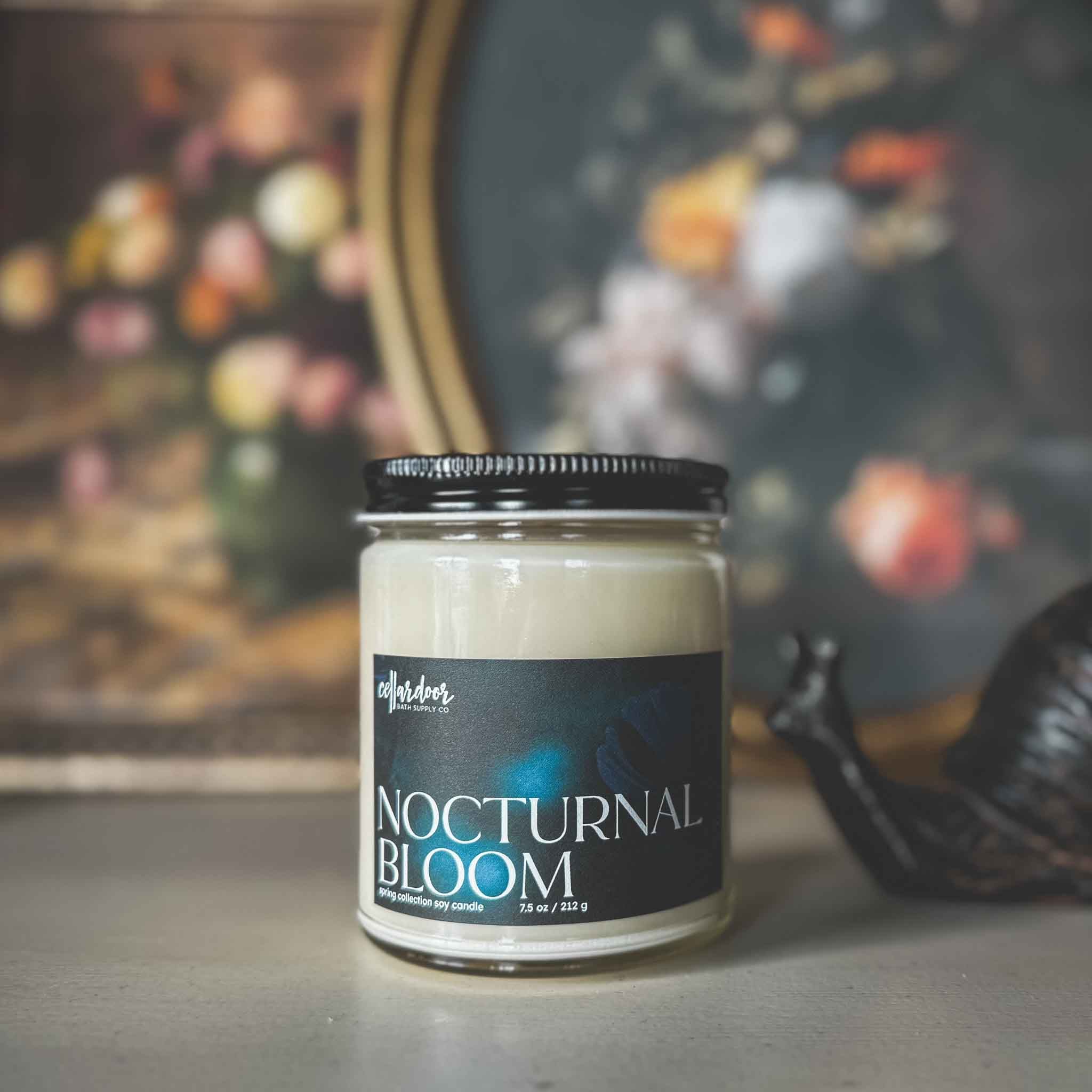 Candle jar labeled 'Nocturnal Bloom' with a blurred painting in the background