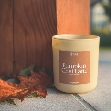 Candle labeled 'Pumpkin Chai Latte' with autumn leaves on a stone surface.