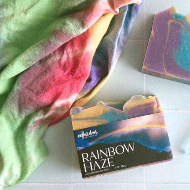 Bar of soap with 'Rainbow Haze' packaging on a white surface, next to a colorful towel.
