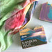 Bar of soap with 'Rainbow Haze' packaging on a white surface, next to a colorful towel.